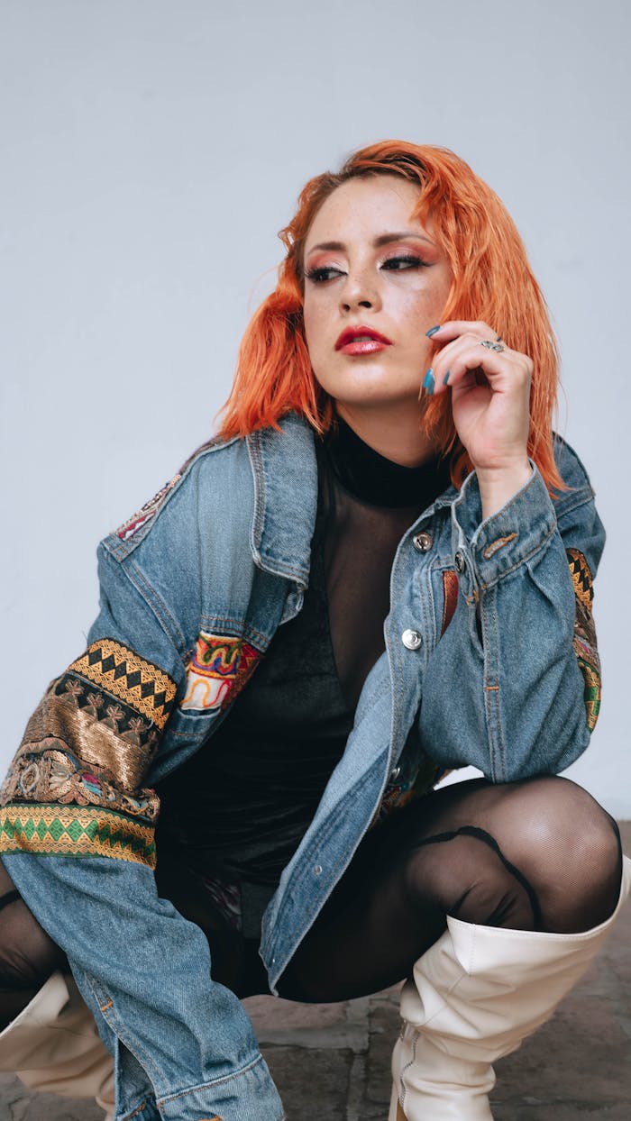 Trendy female model in a vibrant denim jacket and white boots posing indoors.