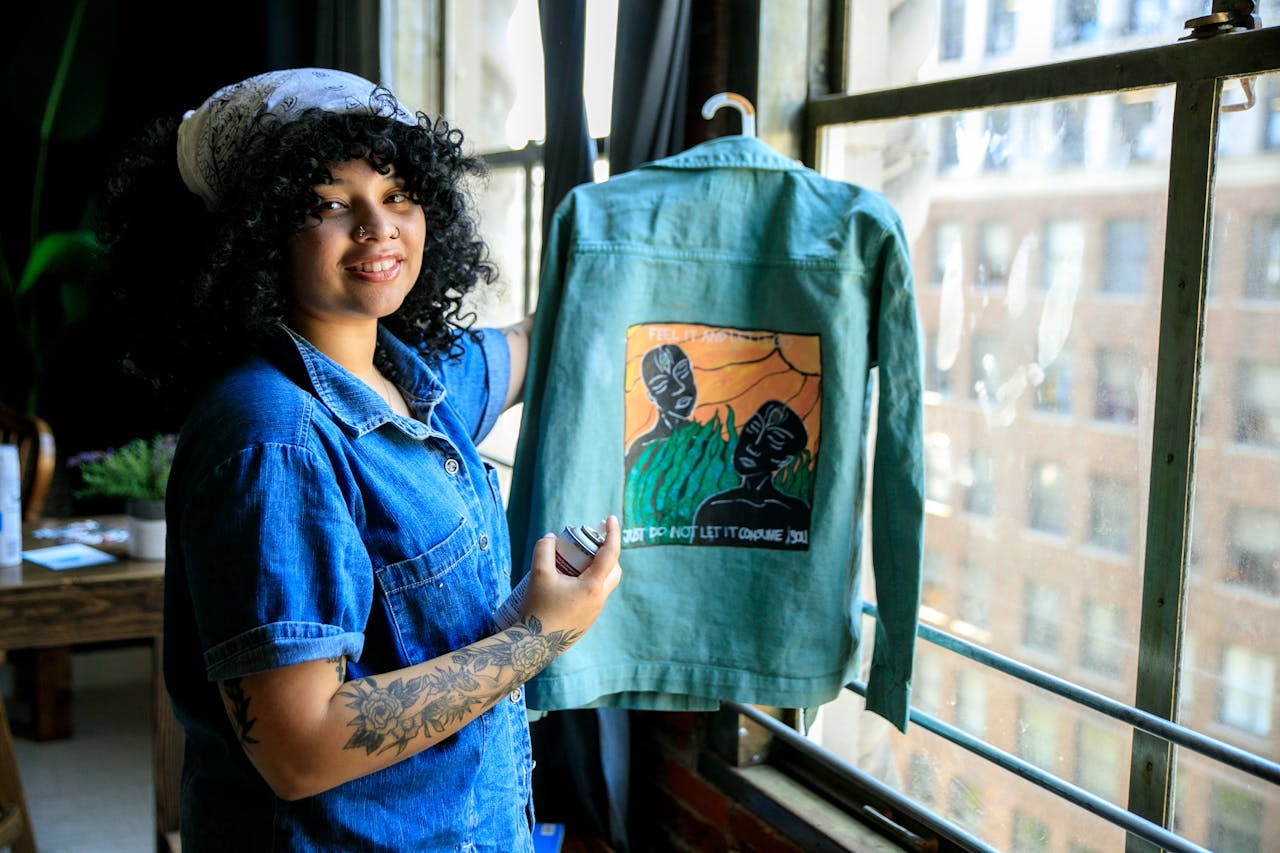 Smiling woman with tattoos displays a custom painted jacket by a window indoors.